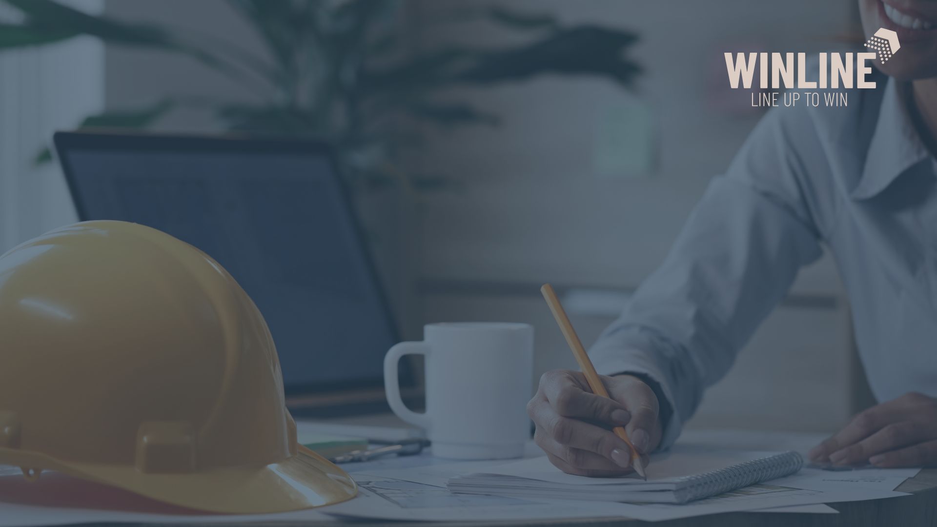 Biurko inżyniera z żółtym kaskiem, laptopem i notatnikiem, osoba w koszuli podpisuje dokumenty przetargowe, w prawym górnym rogu logo Winline